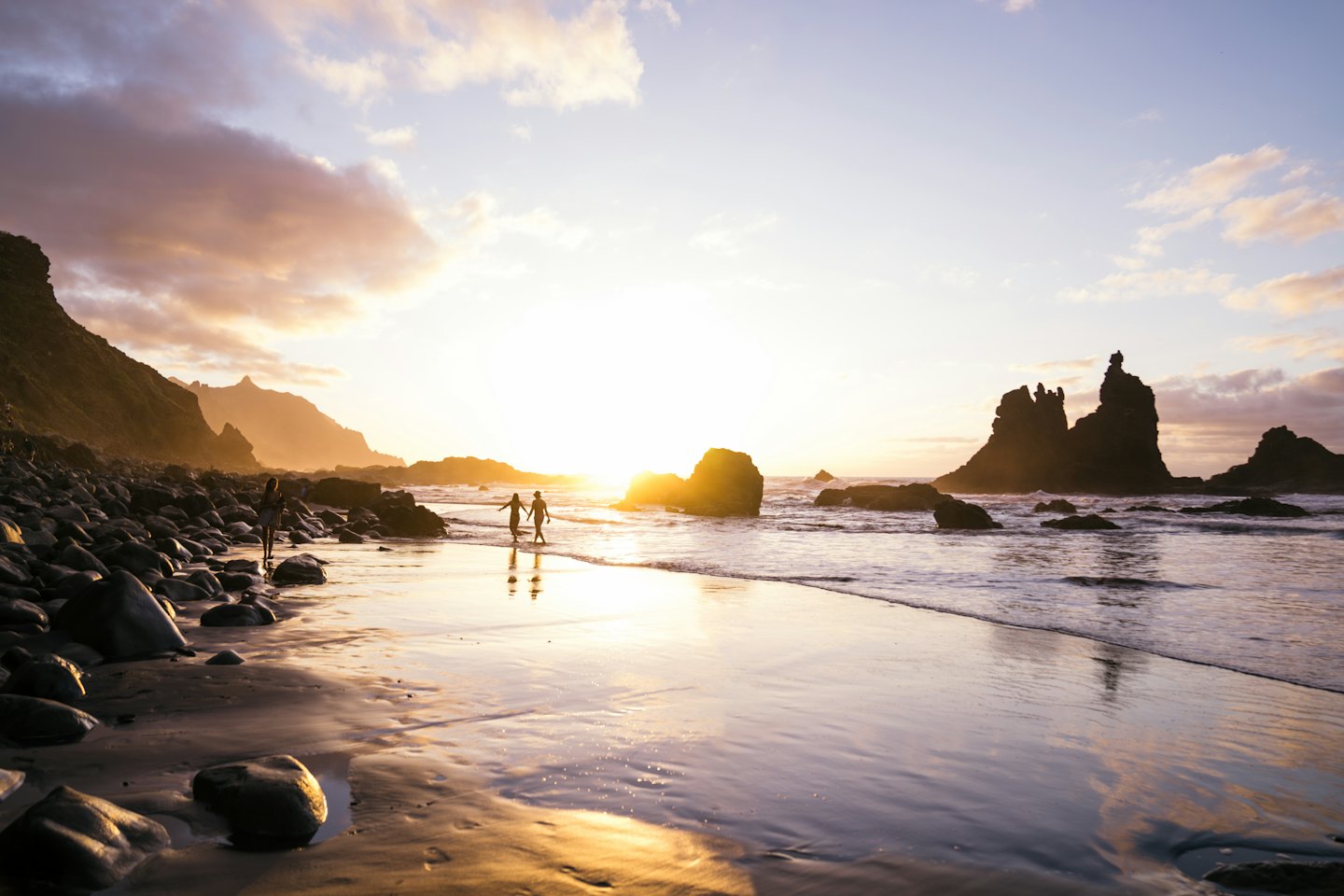 Three people walk along a wet rocky beach at sunset.
