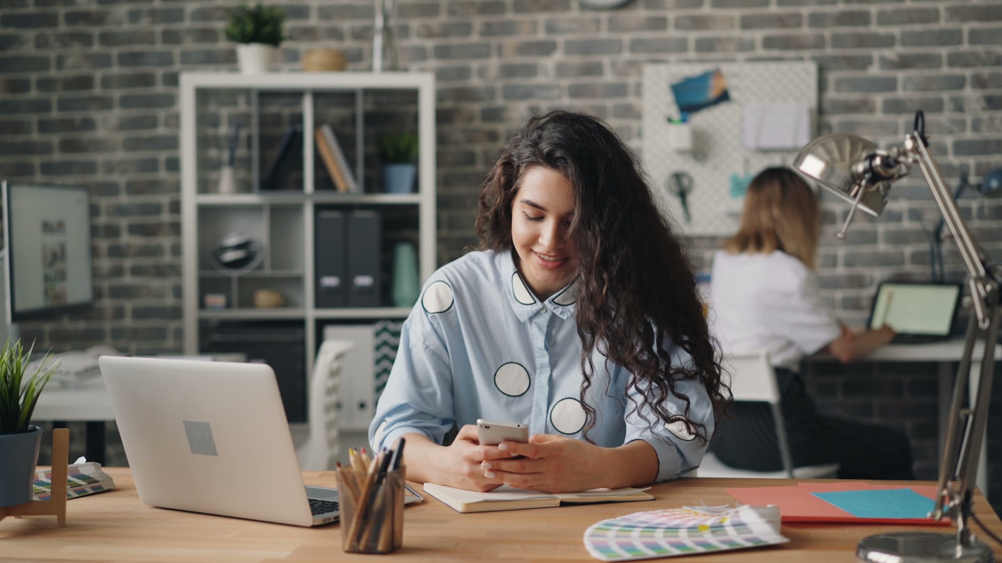 Woman smiles while using her smartphone at a wooden desk.